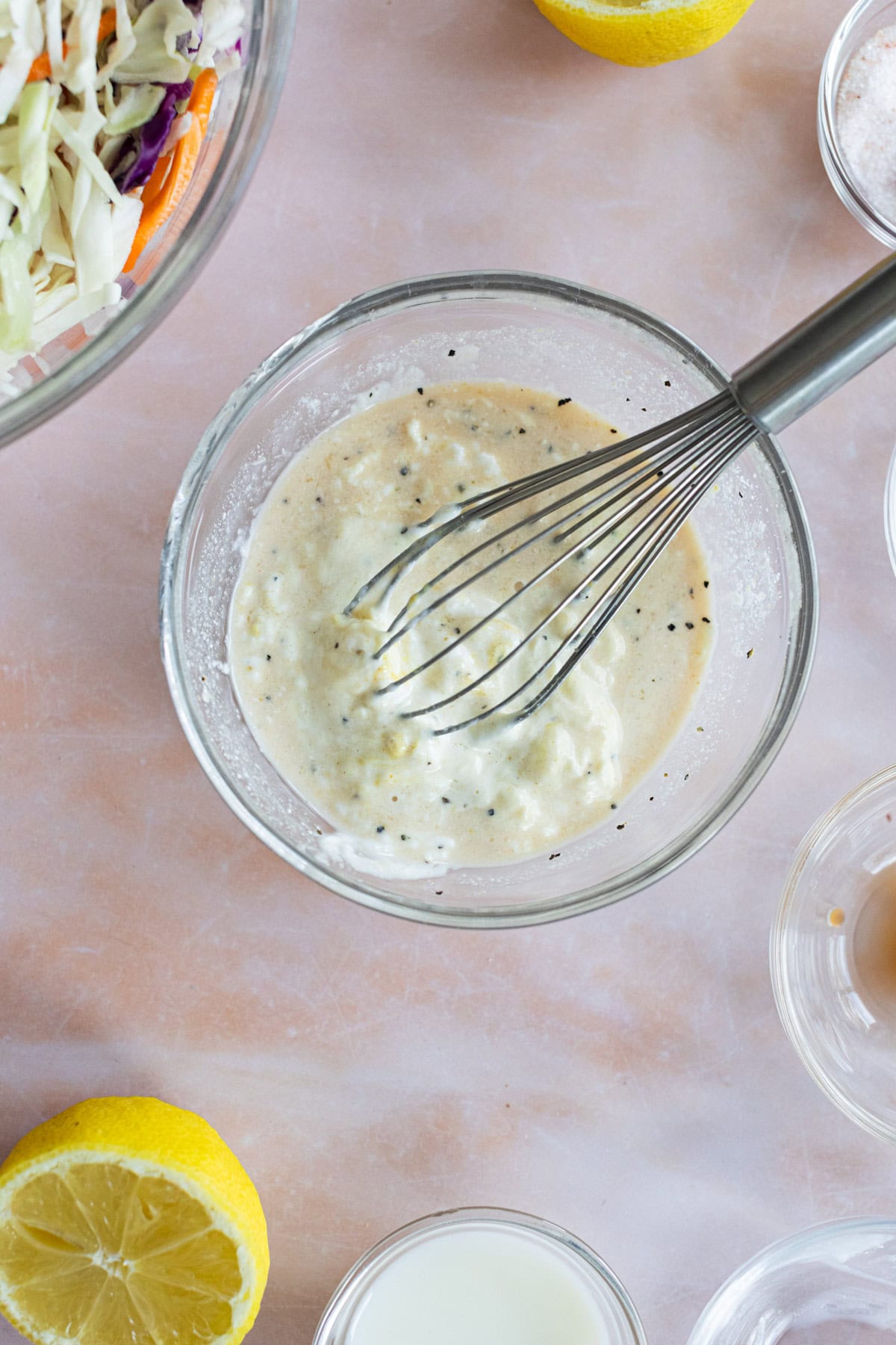 A glass bowl with creamy dressing being whisked, surrounded by lemon halves, cabbage, and small bowls of ingredients on a light surface.