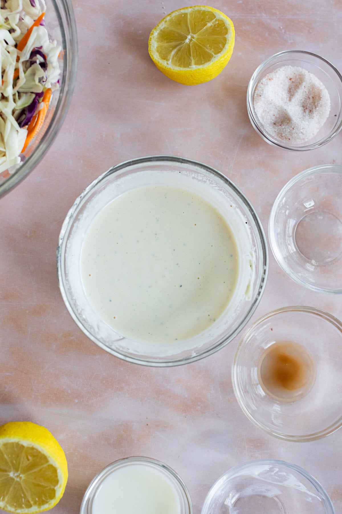 A bowl of creamy dressing is surrounded by lemon halves, seasoning, and empty glass bowls on a light surface. Part of a bowl of shredded salad is visible in the corner.