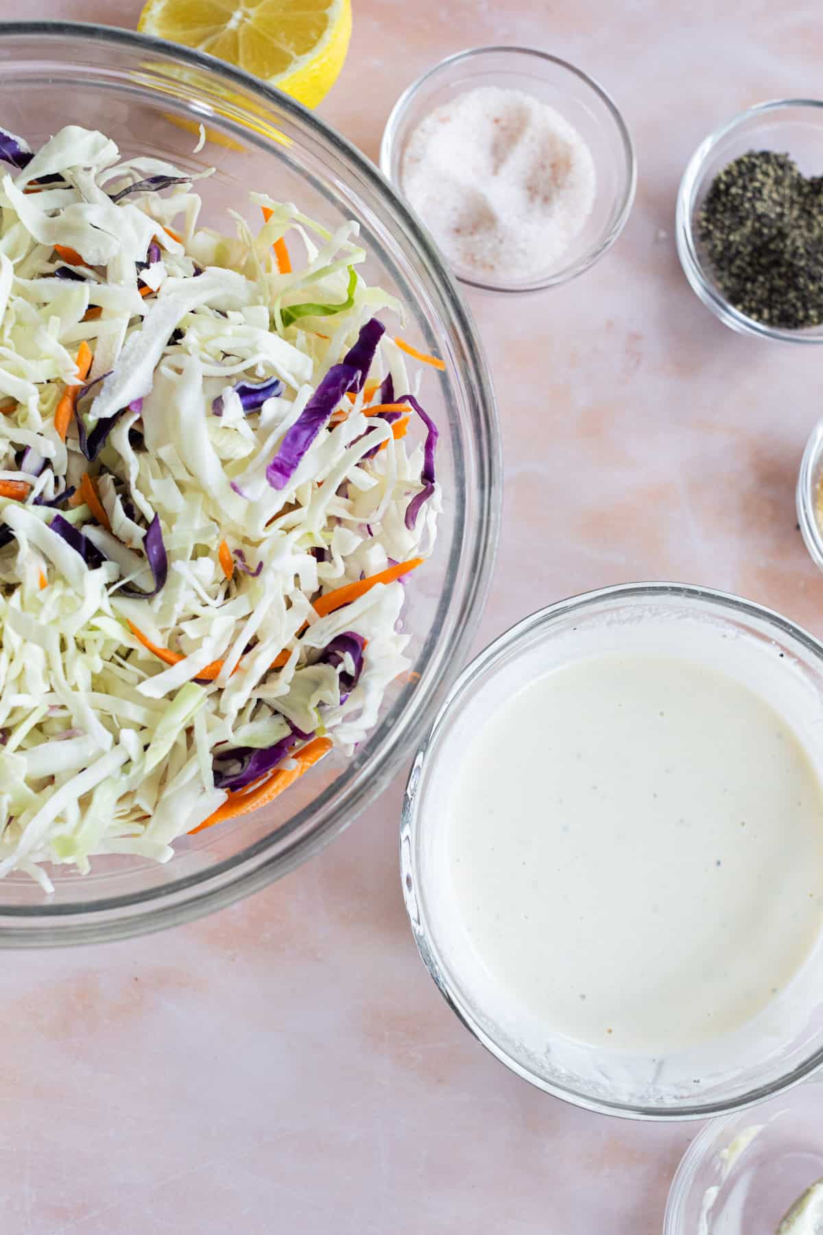 A bowl of shredded cabbage slaw, a bowl of creamy dressing, and small bowls of salt, pepper, and half a lemon on a light surface.
