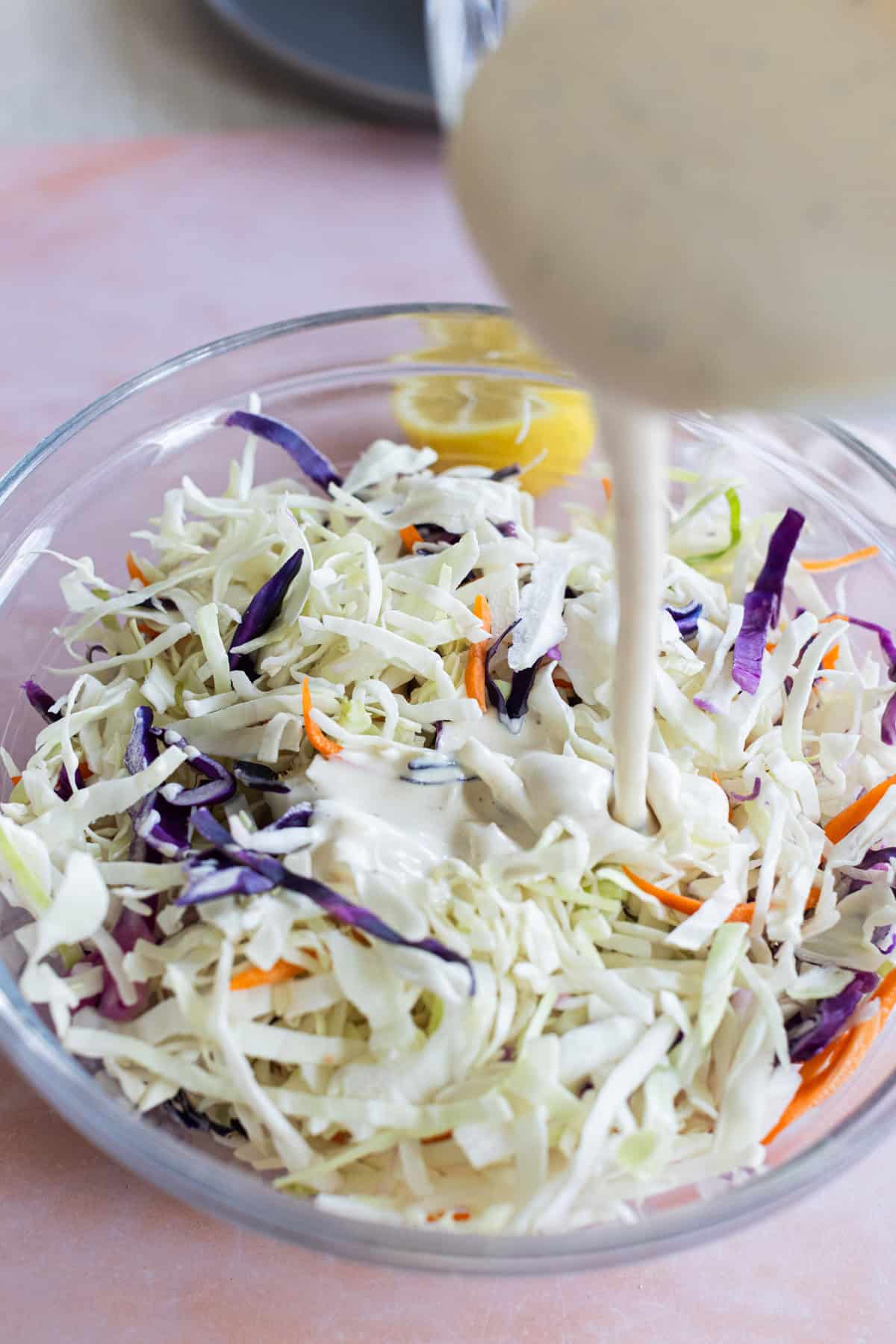 A bowl of shredded cabbage, red cabbage, and carrots with creamy dressing being poured on top.