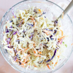 A glass bowl filled with creamy coleslaw made of shredded cabbage, carrots, and purple cabbage, with a serving spoon and lemon halves nearby.