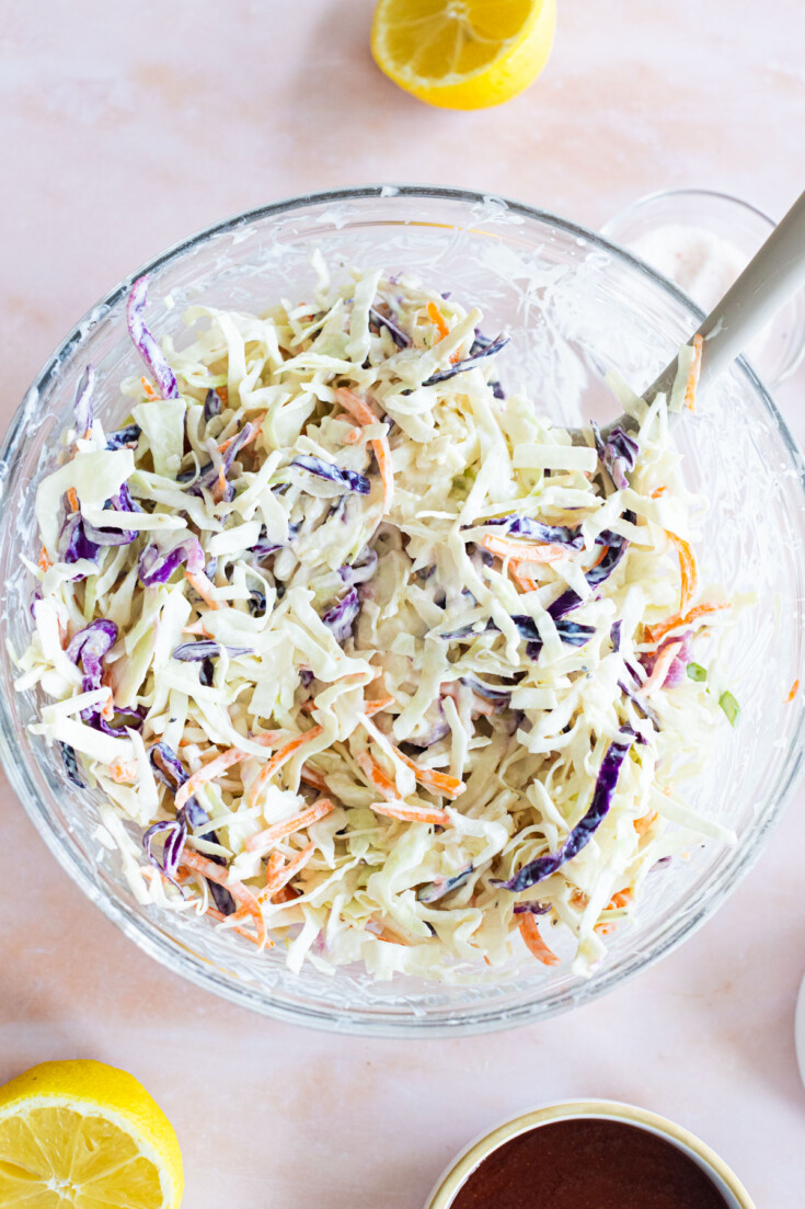 A glass bowl filled with creamy coleslaw made of shredded cabbage, carrots, and purple cabbage, with a serving spoon and lemon halves nearby.