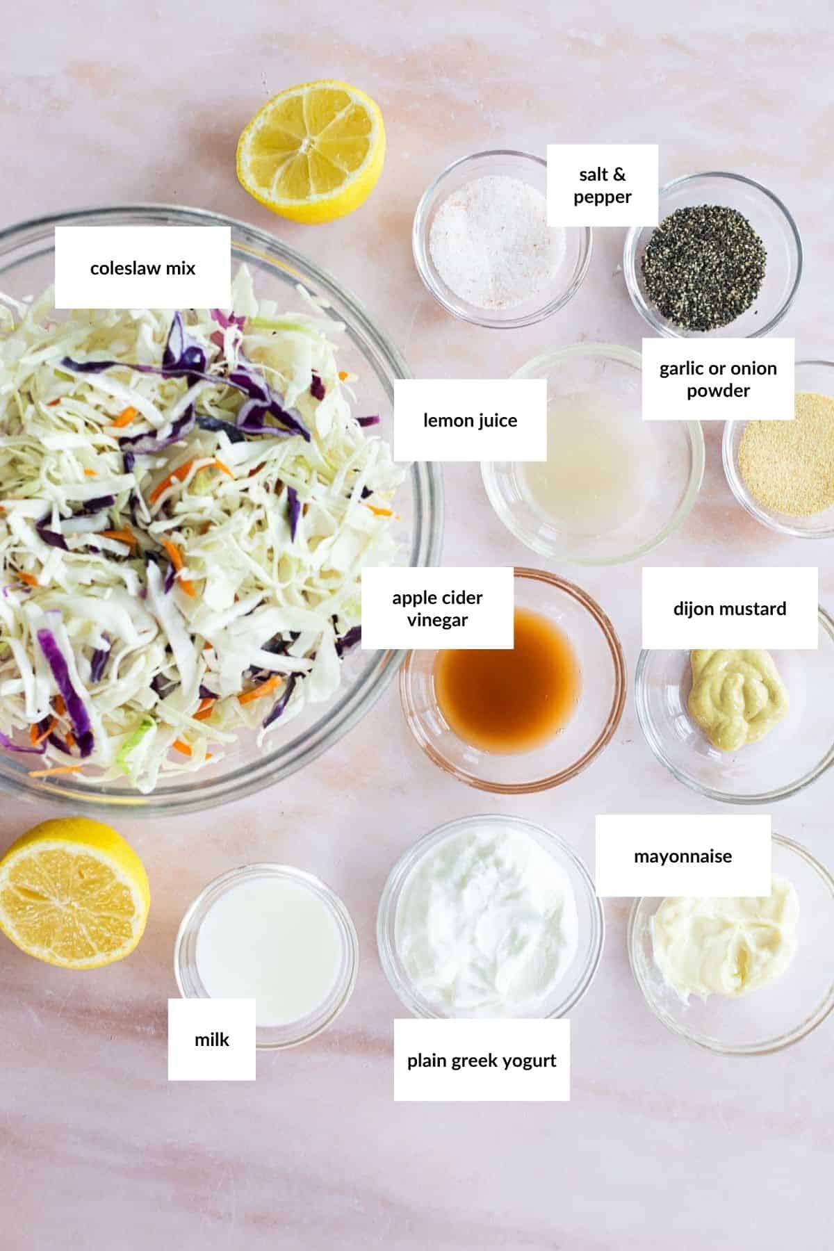 A bowl of coleslaw mix surrounded by small bowls containing lemon juice, apple cider vinegar, dijon mustard, mayonnaise, plain Greek yogurt, milk, salt, pepper, and garlic or onion powder.
