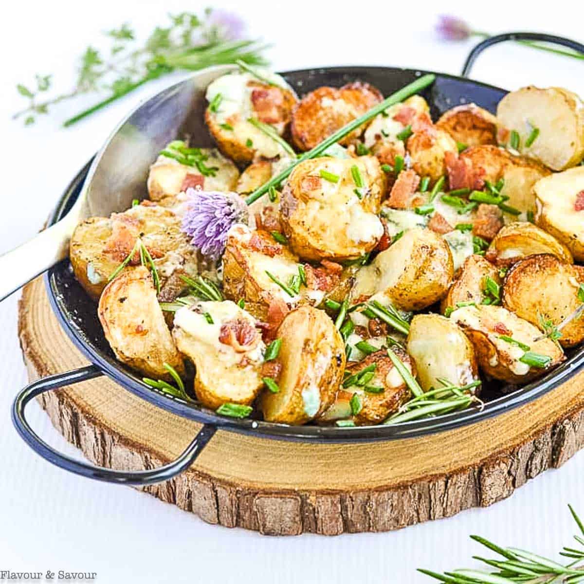 A skillet of roasted baby potatoes topped with melted cheese, bacon, and chopped herbs, placed on a wooden serving board.