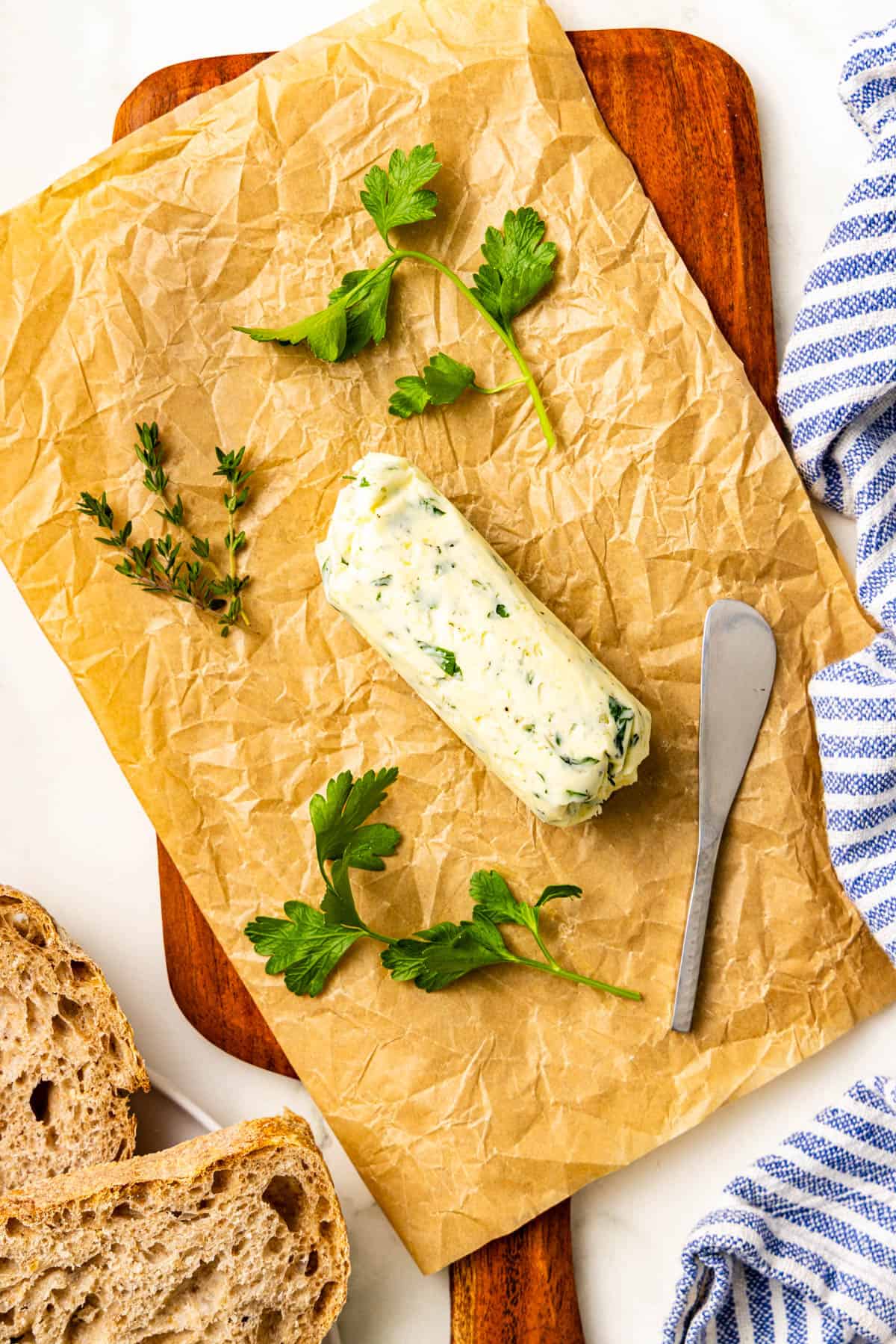 A log of herb butter on parchment paper with parsley, thyme, a butter knife, and slices of bread on a wooden board.