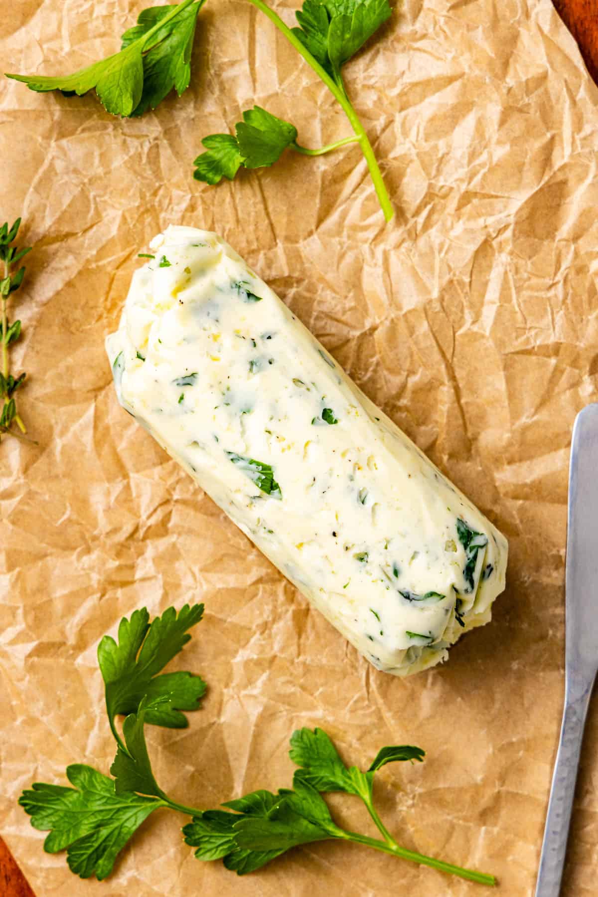 A log of herb butter with visible green herbs sits on crumpled parchment paper, surrounded by fresh parsley leaves and a knife.