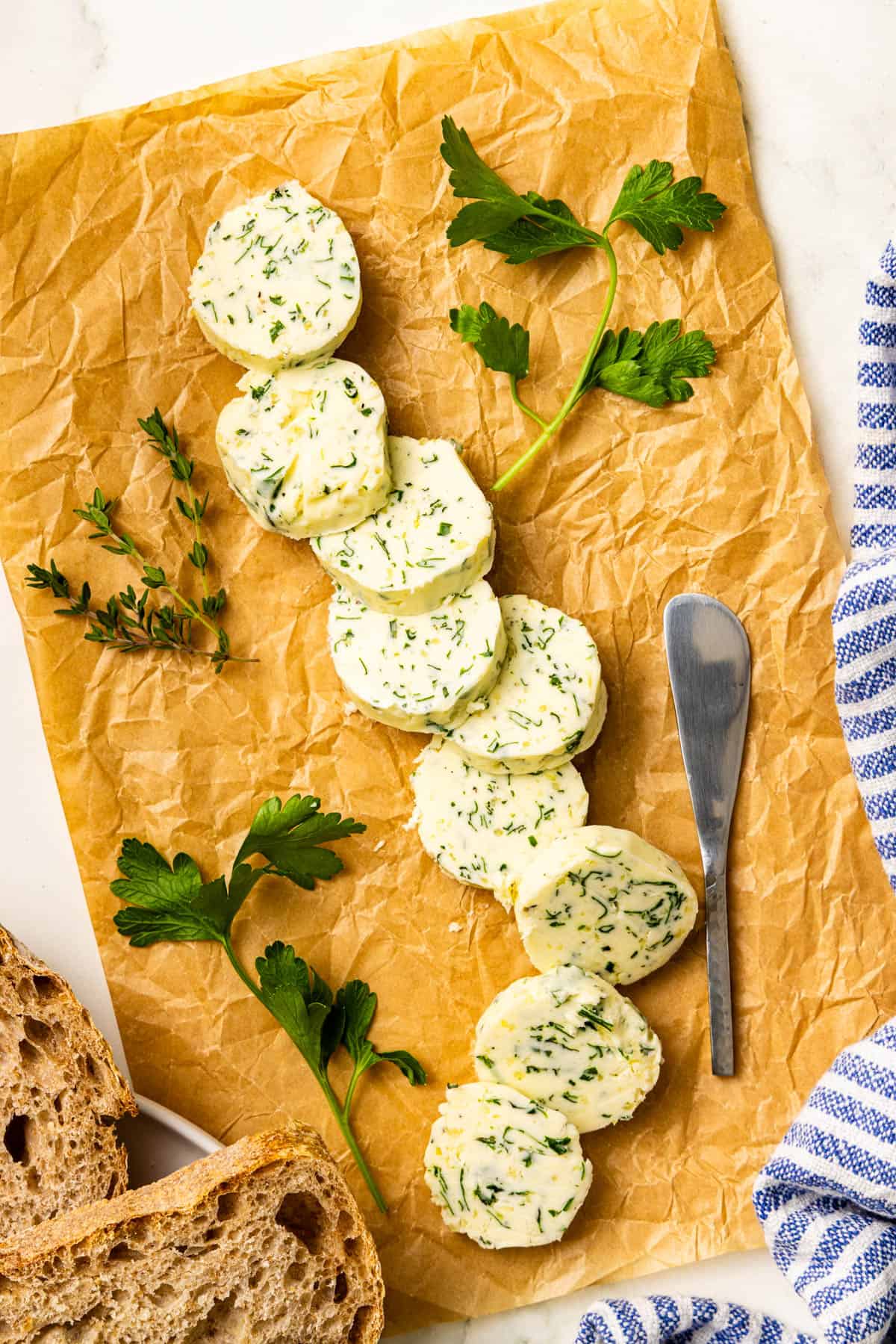 Sliced herb butter arranged on brown parchment paper with fresh herbs, a butter knife, and slices of rustic bread nearby.