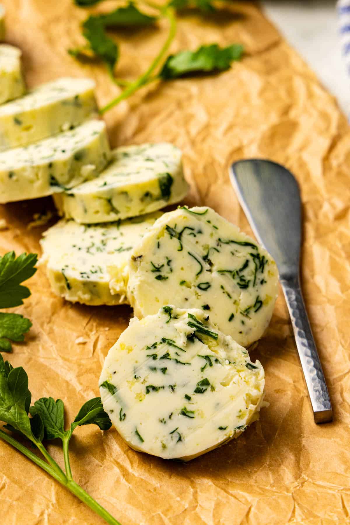 Sliced compound butter with herbs displayed on parchment paper, garnished with fresh parsley, alongside a butter knife.