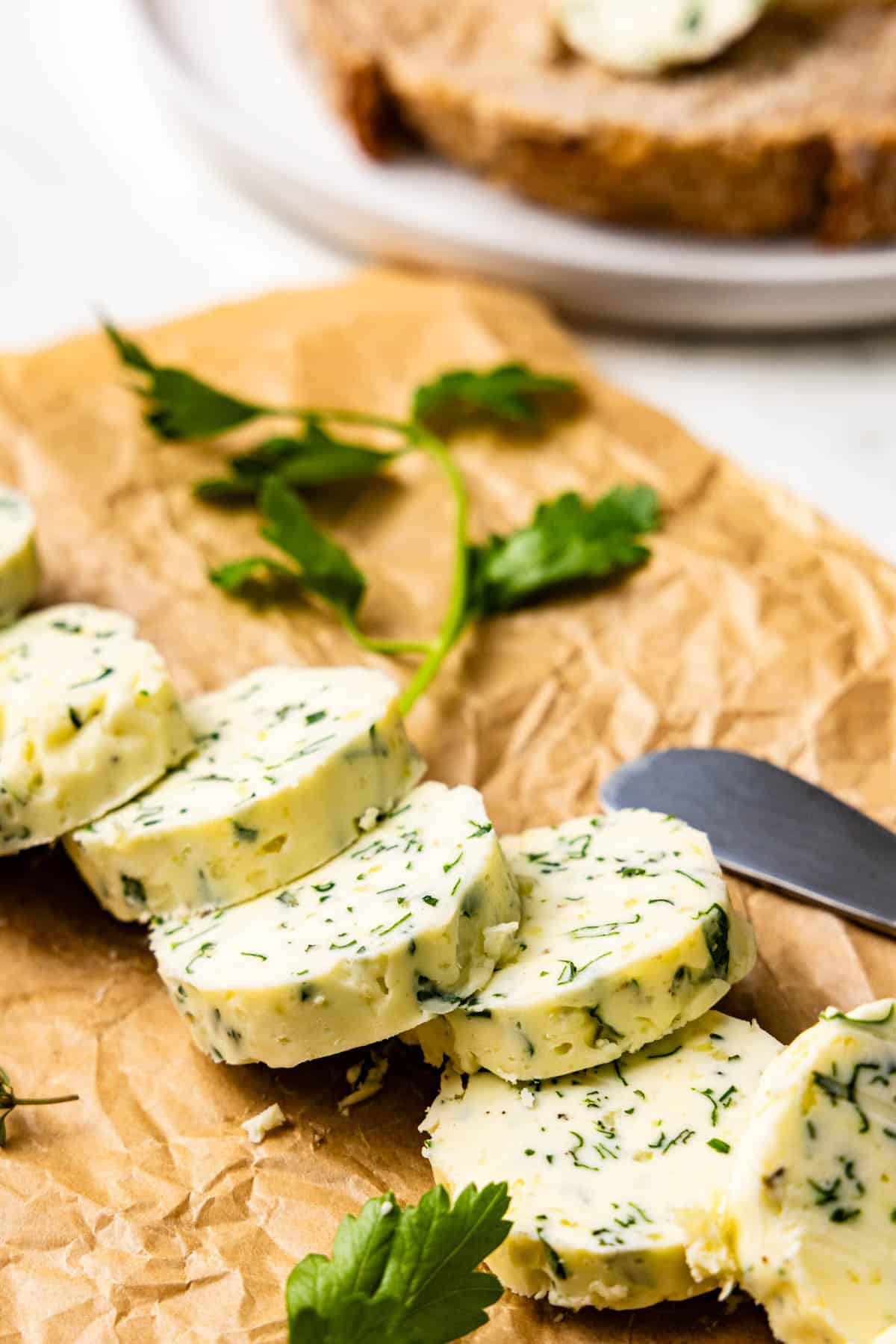 Sliced herb butter on parchment paper with a knife and parsley garnish, with a slice of bread in the background.