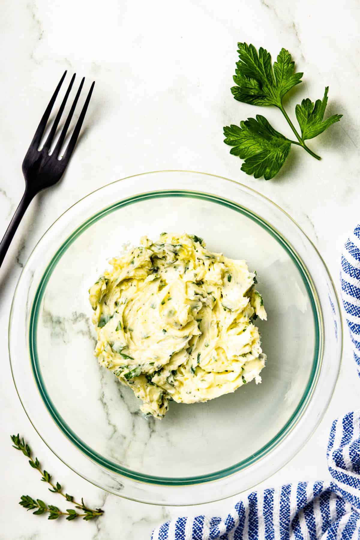 A glass bowl with herb butter on a marble surface, next to a fork, fresh herbs, and a blue-striped cloth.