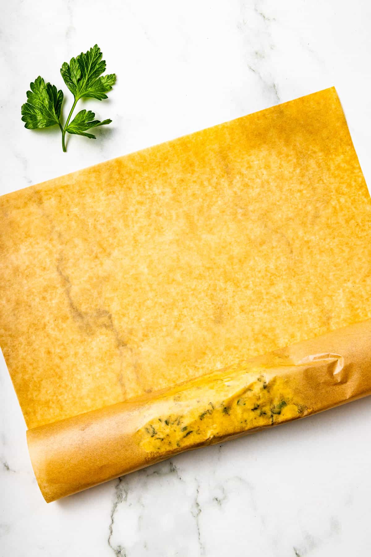 A sheet of parchment paper partially rolled up with a herb mixture inside, placed on a white marble surface next to two parsley leaves.