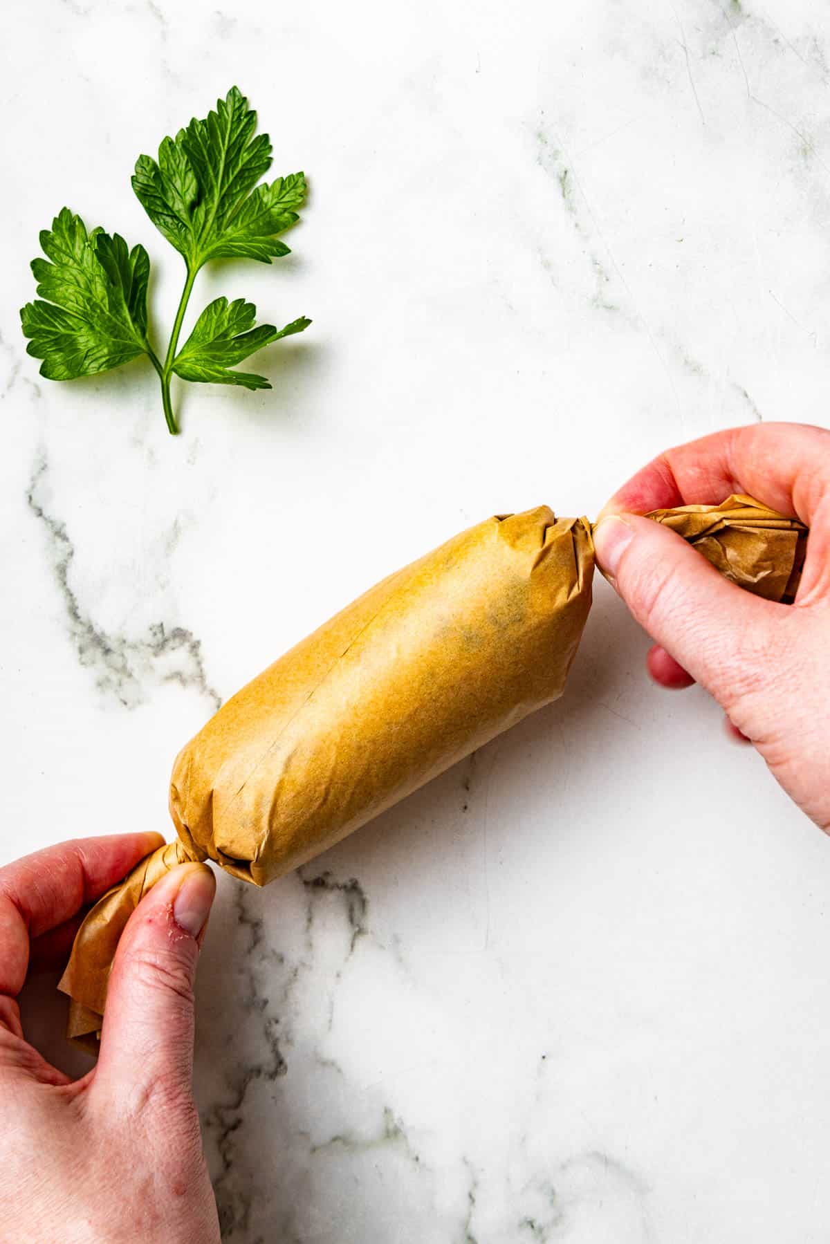 Two hands twist the ends of a brown parchment paper-wrapped item on a white marble surface, with a sprig of green parsley nearby.