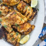 Grilled chicken thighs garnished with chopped herbs and served with lime wedges on a white oval plate.