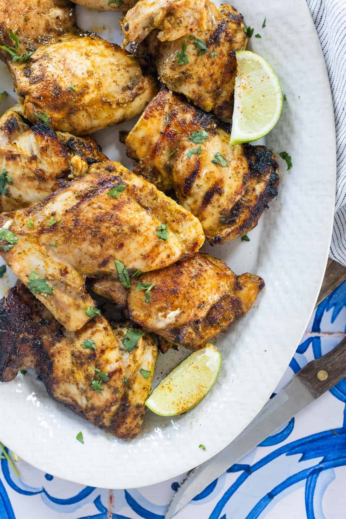 Grilled chicken thighs garnished with chopped herbs and served with lime wedges on a white oval plate.