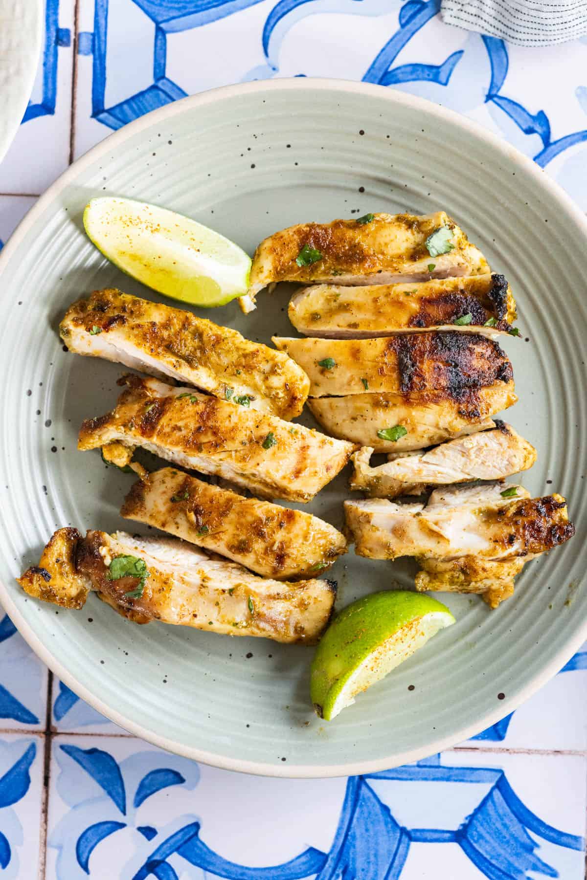 Sliced grilled chicken breast on a green plate, garnished with herbs and served with two lime wedges on a blue and white patterned surface.