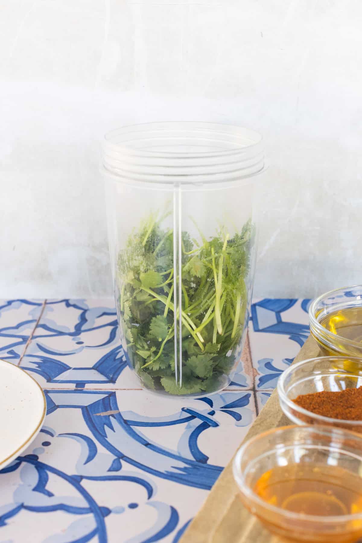 A clear blender cup filled with fresh cilantro sits on a patterned blue and white surface, next to small bowls containing spices and liquids.