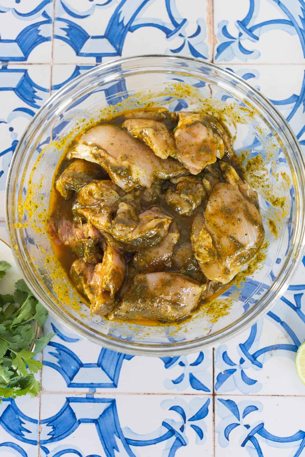 Raw chicken pieces marinating in Mexican chicken marinade in a glass bowl on a blue and white tiled surface, with cilantro and a lime slice nearby.