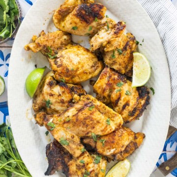 Grilled chicken thighs garnished with chopped herbs and lime wedges, served on a white oval plate with a blue and white patterned background.