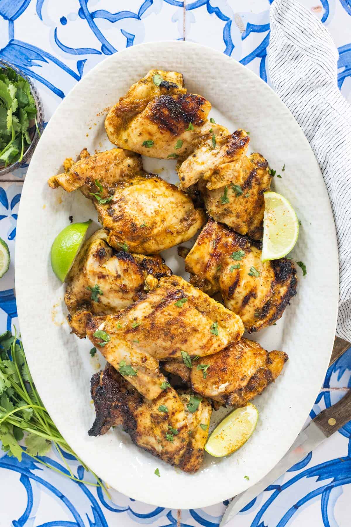 Grilled chicken thighs garnished with chopped herbs and lime wedges, served on a white oval plate with a blue and white patterned background.
