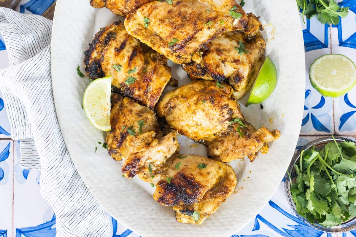 Grilled chicken thighs garnished with chopped herbs and lime wedges on a white oval plate, set on a blue and white tile surface.