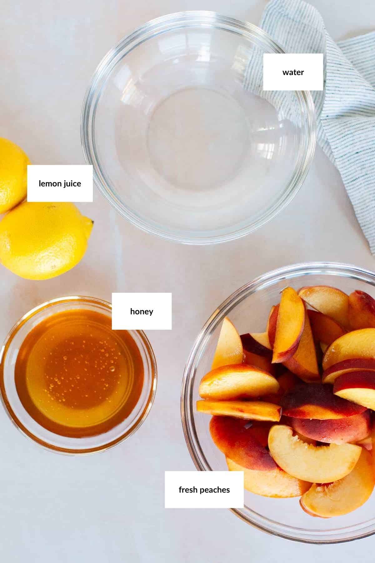 Top-down view of bowls containing water, honey, and sliced fresh peaches, with whole lemons and a small bowl of lemon juice on a white surface.