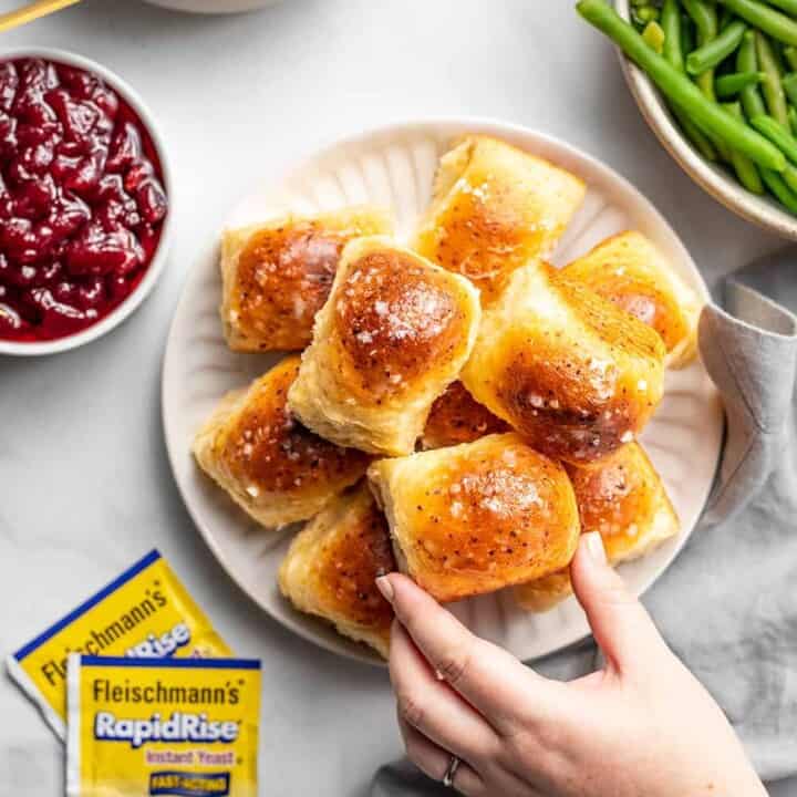 A hand reaching for a golden brown dinner roll on a plate, with packets of yeast, cranberry sauce, and green beans nearby.