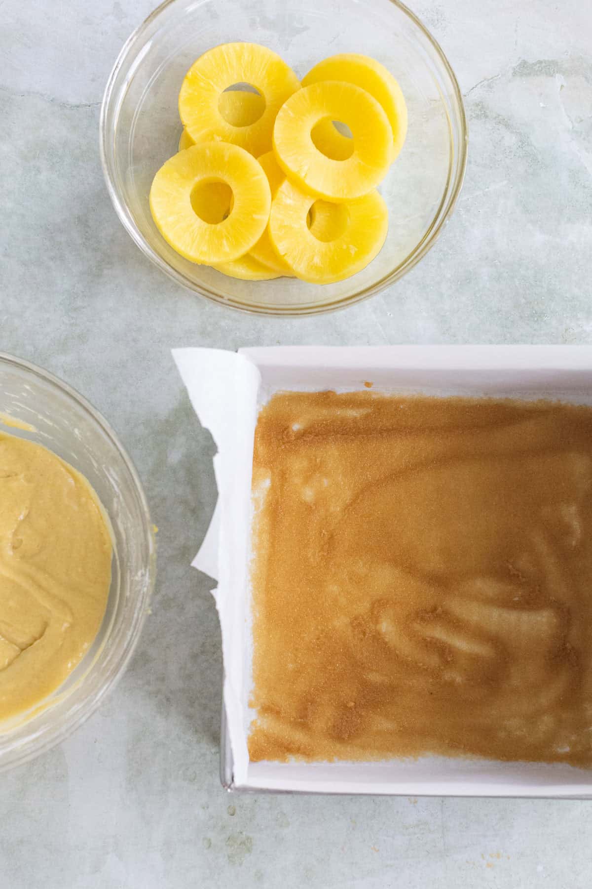 A bowl of pineapple rings, a bowl of cake batter, and a parchment-lined baking pan with a brown sugar layer are arranged on a light surface.