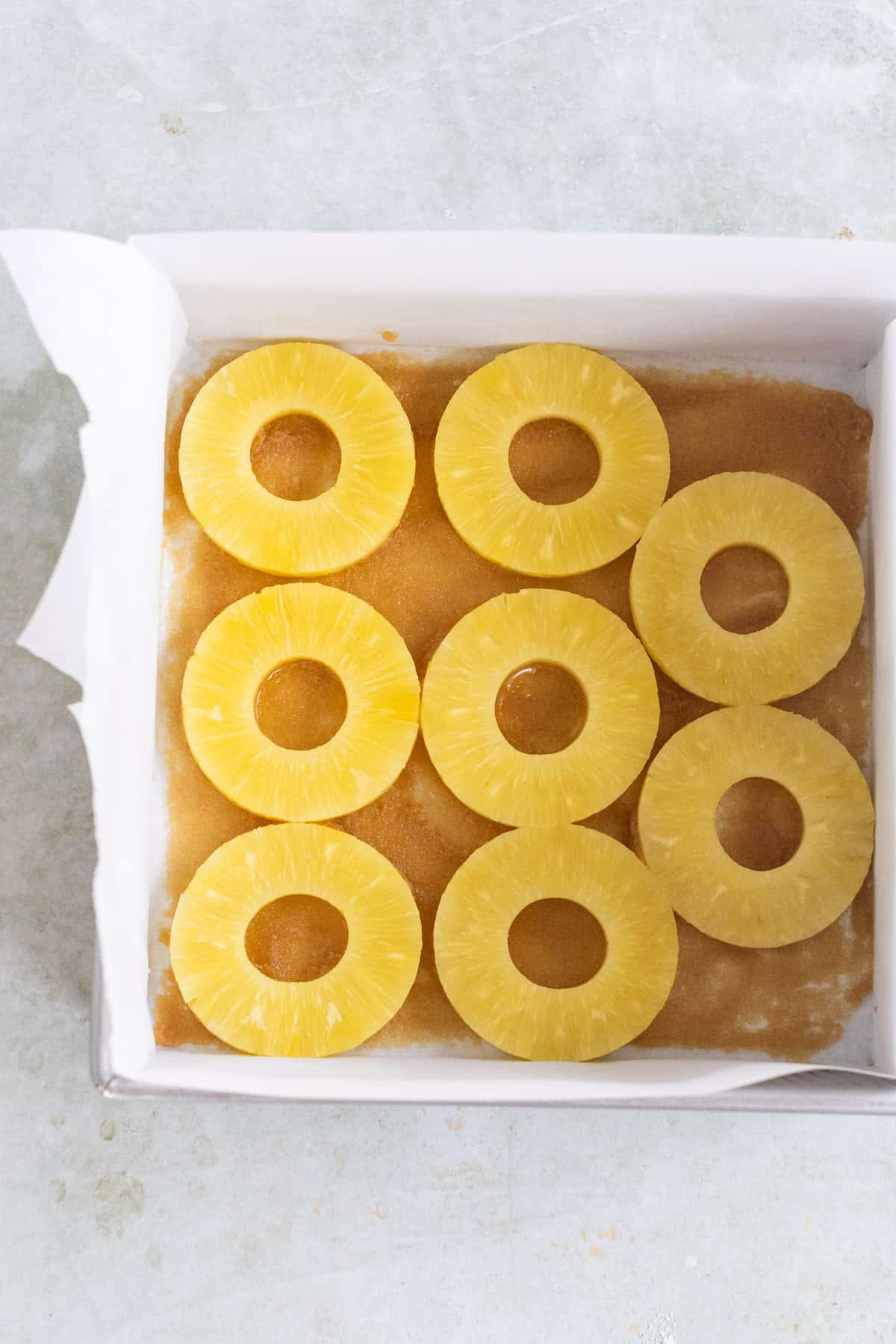 A square baking dish lined with parchment paper, containing nine pineapple rings arranged on top of brown cake batter.