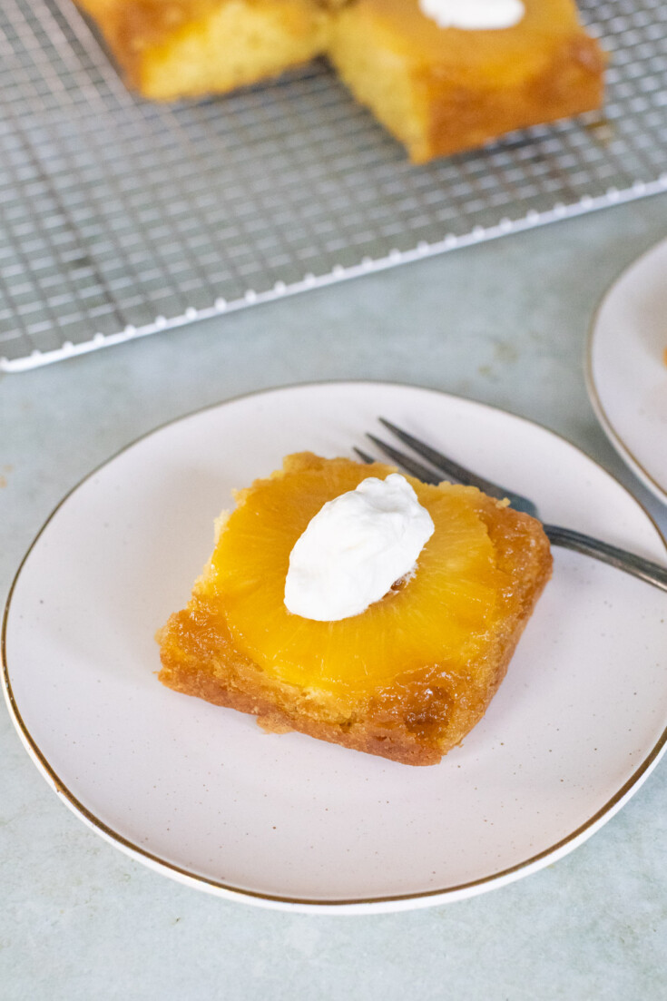 A square slice of pineapple upside-down cake with a dollop of whipped cream on top sits on a plate with a fork. More cake is visible on a cooling rack in the background.