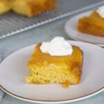 A square piece of yellow pineapple upside-down cake topped with a dollop of whipped cream on a white plate, with more cake pieces in the background.
