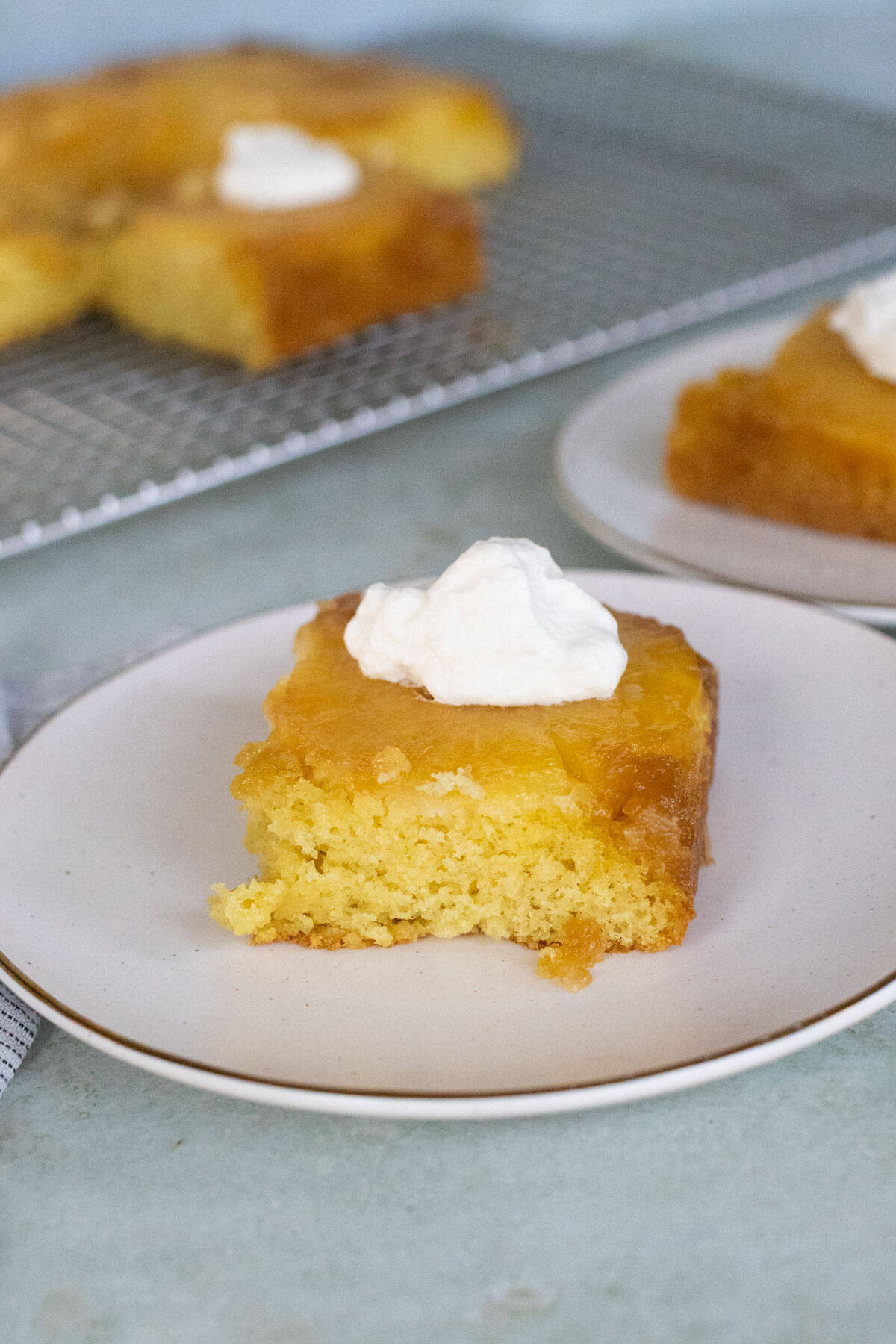 A square piece of yellow pineapple upside-down cake topped with a dollop of whipped cream on a white plate, with more cake pieces in the background.