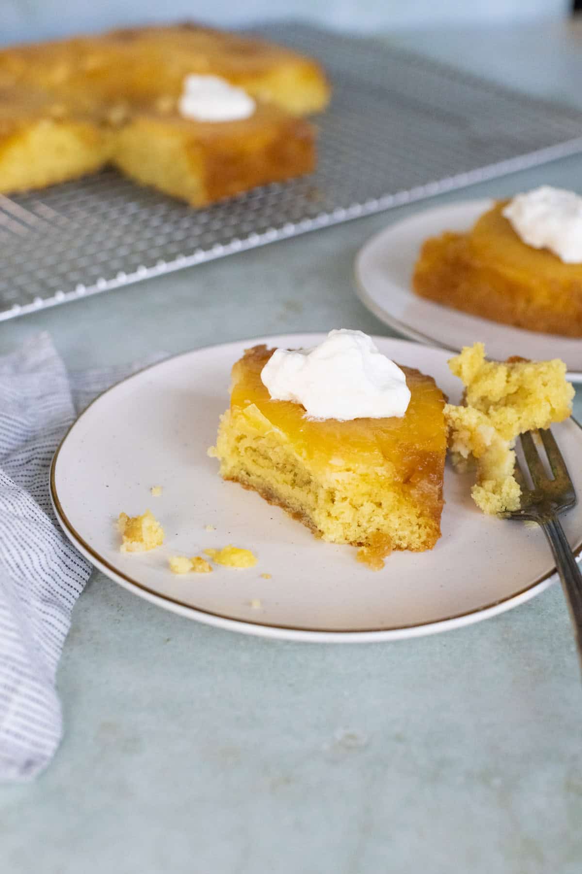 A slice of yellow cake with a layer of fruit topping and whipped cream on a plate, with a fork and additional cake slices in the background.