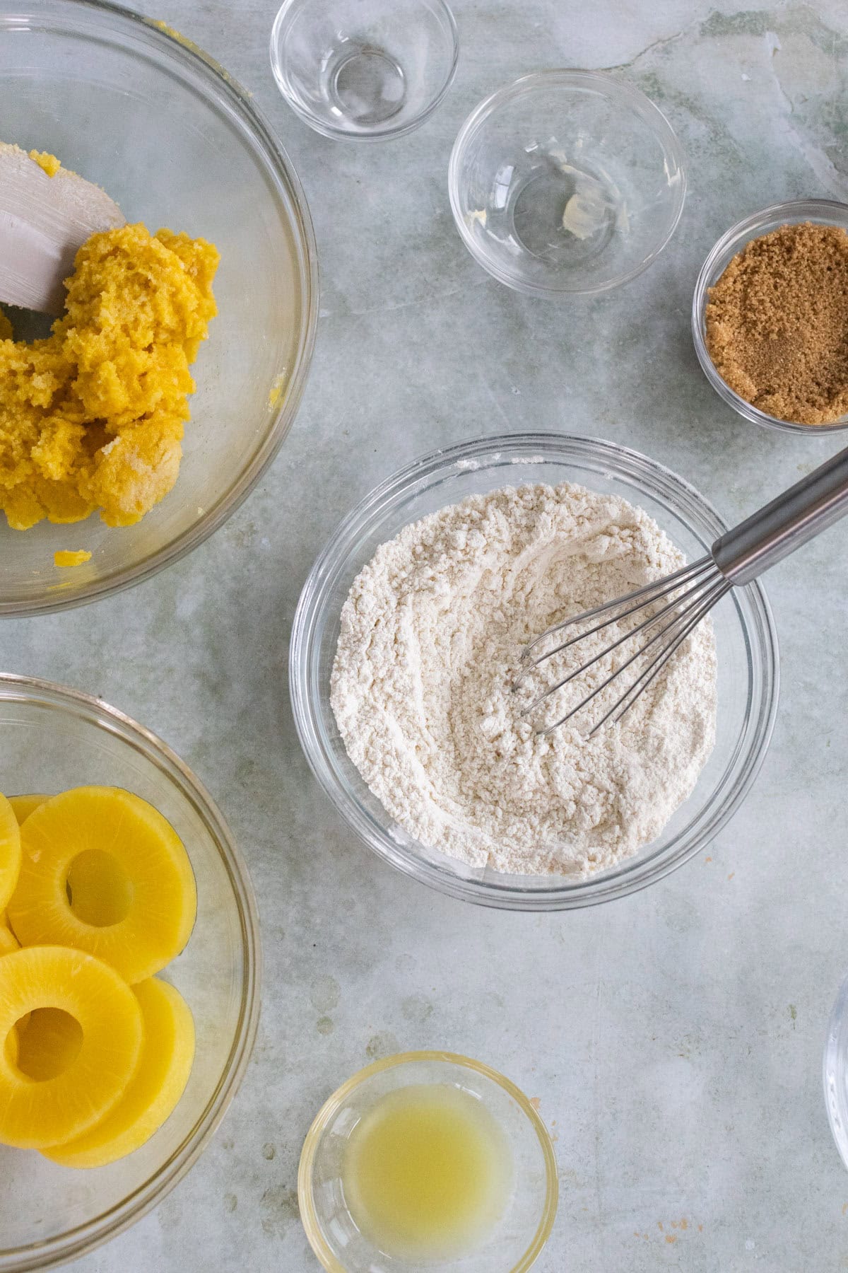 Bowls with flour and a whisk, brown sugar, pineapple rings, lemon juice, and a yellow batter are arranged on a light countertop.