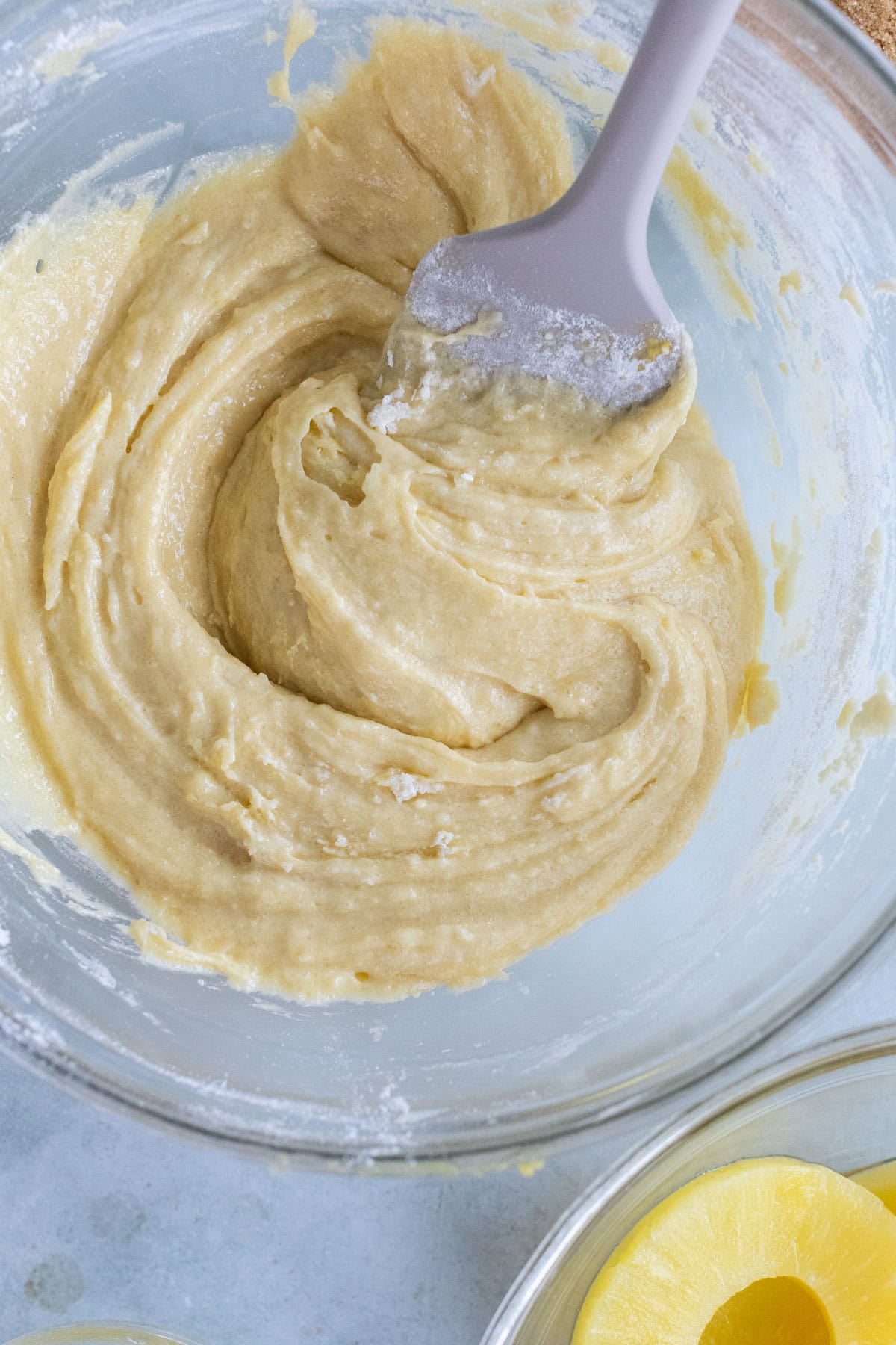 A mixing bowl with thick yellow cake batter being stirred by a gray spatula. Pineapple slices are visible nearby.
