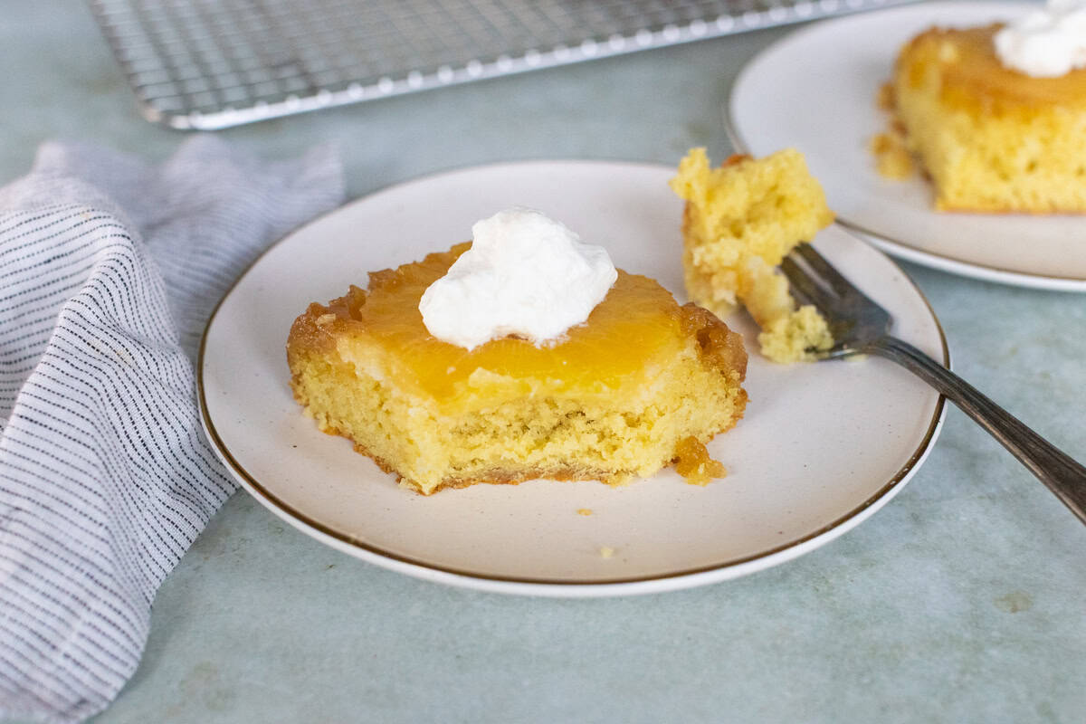 A slice of yellow cake topped with a dollop of whipped cream and fruit glaze sits on a white plate with a fork and a striped cloth nearby.