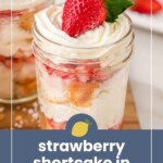 A mason jar filled with layers of whipped cream, strawberries, and cake, topped with a whole strawberry. Text reads "strawberry shortcake in cups.