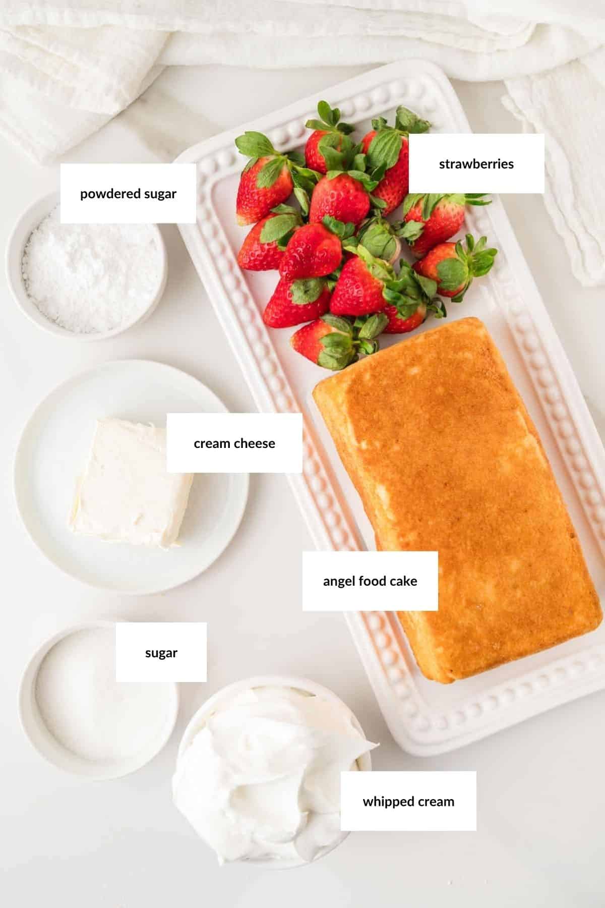 A rectangular platter holds strawberries and angel food cake, with bowls of powdered sugar, cream cheese, sugar, and whipped cream arranged beside it. Labels identify each ingredient.