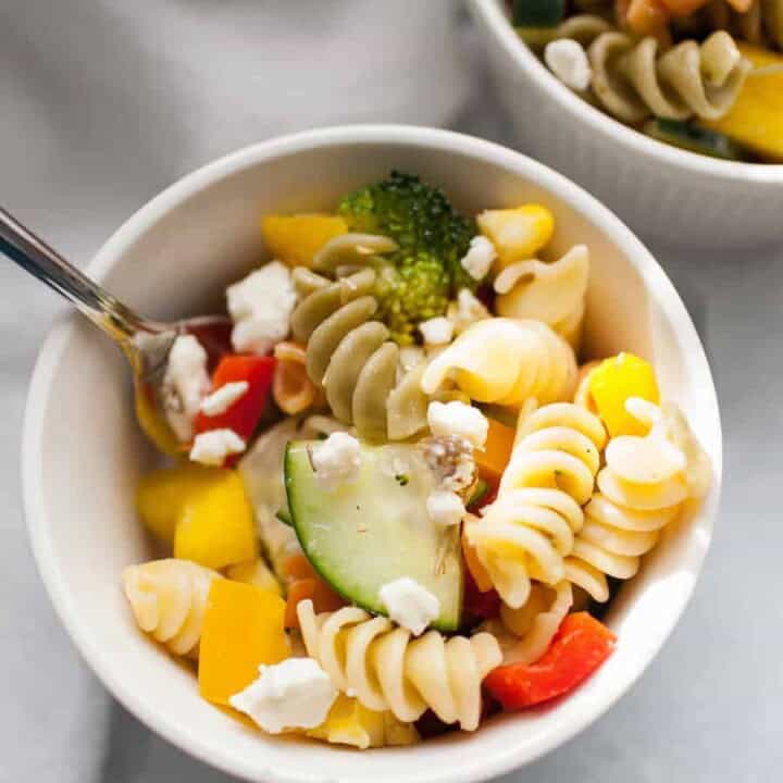 A bowl of rotini pasta salad with broccoli, cucumber, bell peppers, and crumbled feta cheese, with a fork resting inside.