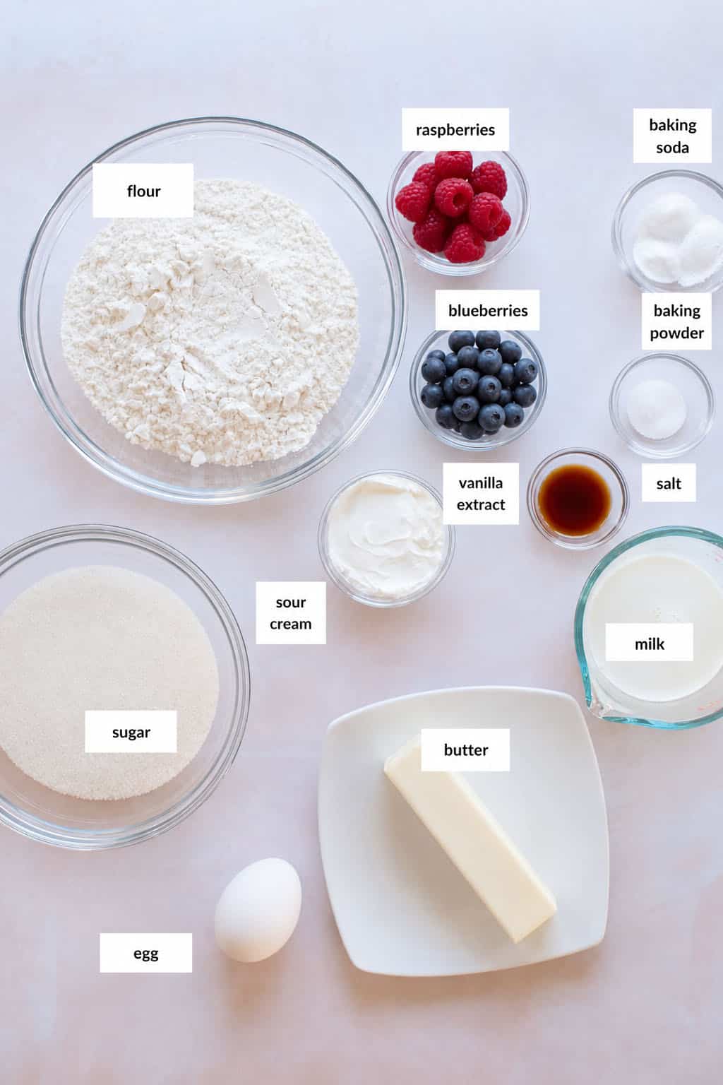 Various labeled baking ingredients, including flour, sugar, sour cream, egg, butter, milk, vanilla extract, raspberries and blueberries—are arranged on a countertop.