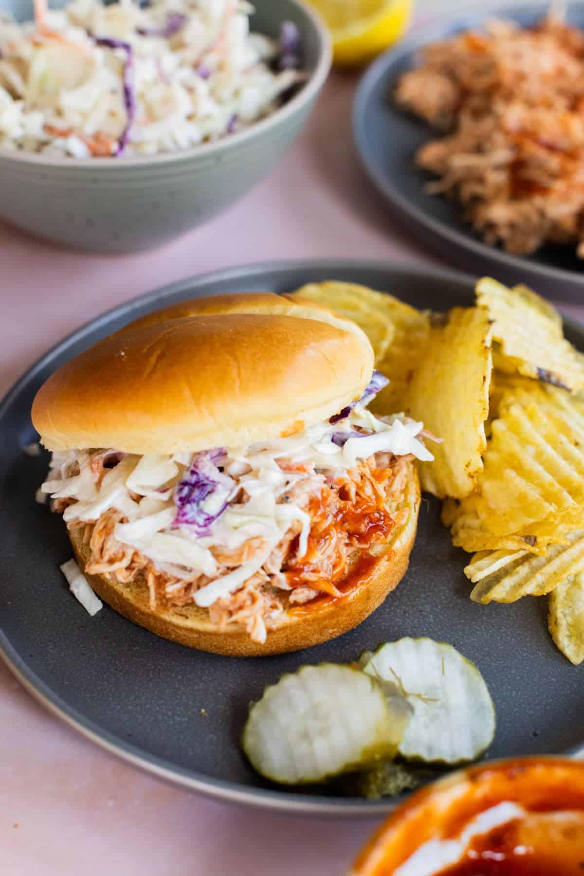 A bbq chicken sandwich with creamy coleslaw on a bun, served with crinkle-cut potato chips and pickle slices on a gray plate.
