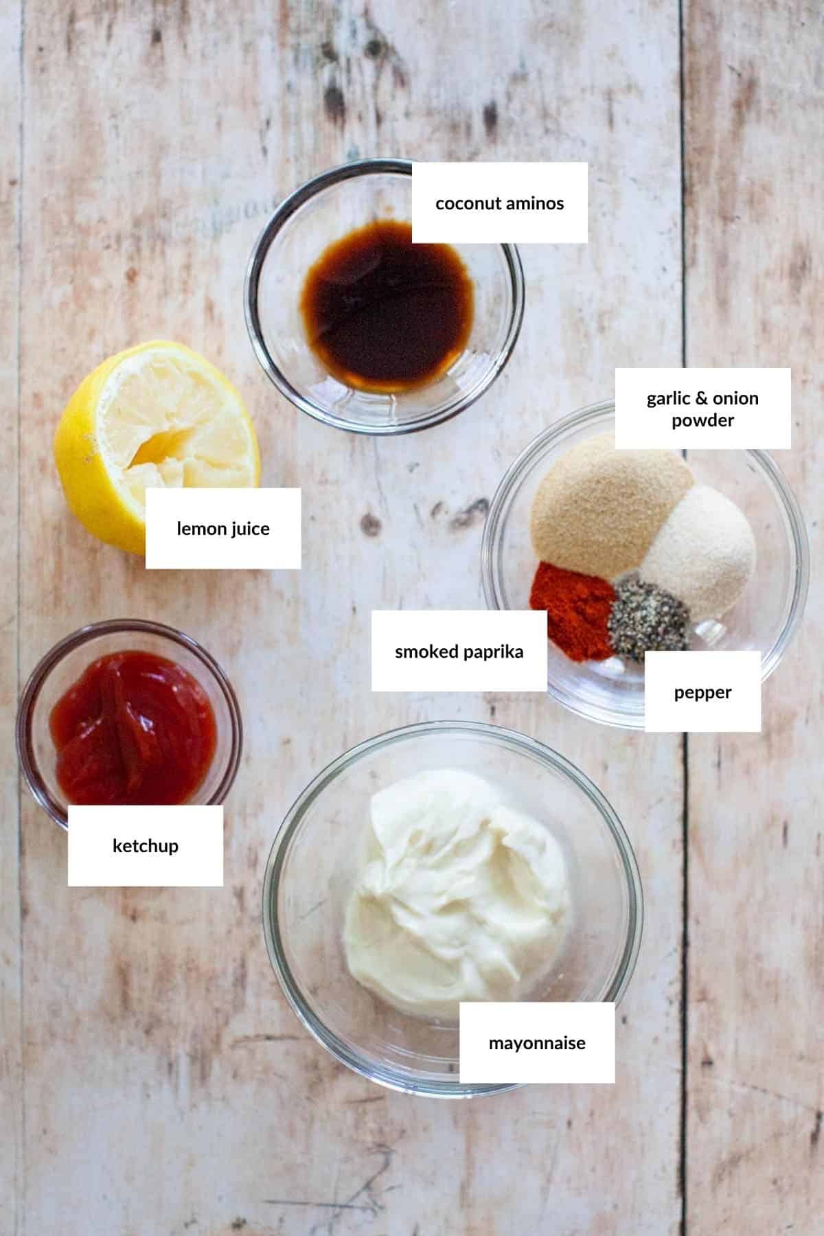 Five small bowls of ingredients and a half lemon on a wooden surface, each labeled: coconut aminos, lemon juice, ketchup, smoked paprika with garlic and onion powder plus pepper, and mayonnaise.