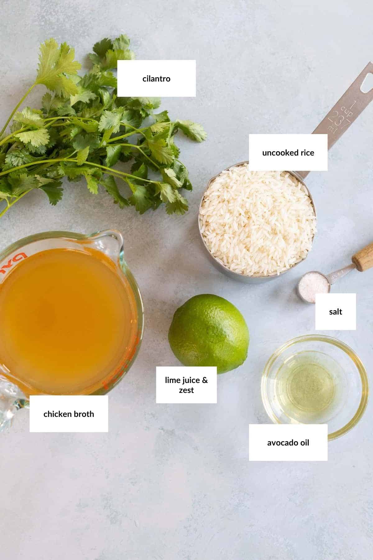 Overhead view of cilantro, uncooked rice, salt, avocado oil, chicken broth, and a lime on a light surface, each ingredient labeled.