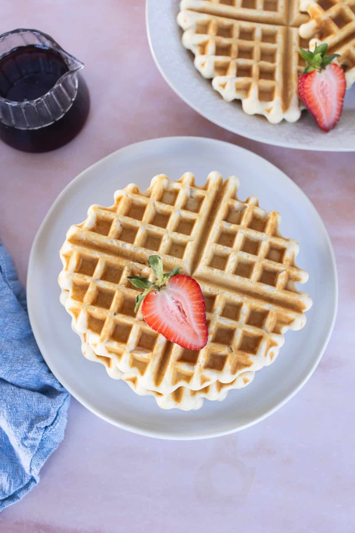 A plate with an Easy Waffles round waffle topped with a halved strawberry, next to a pitcher of syrup and a partial plate of waffles in the background.
