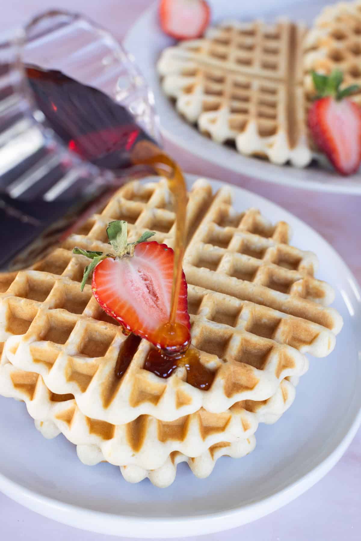 Syrup is being poured onto a stack of plated waffles topped with a sliced strawberry. Another plate of waffles sits in the background.