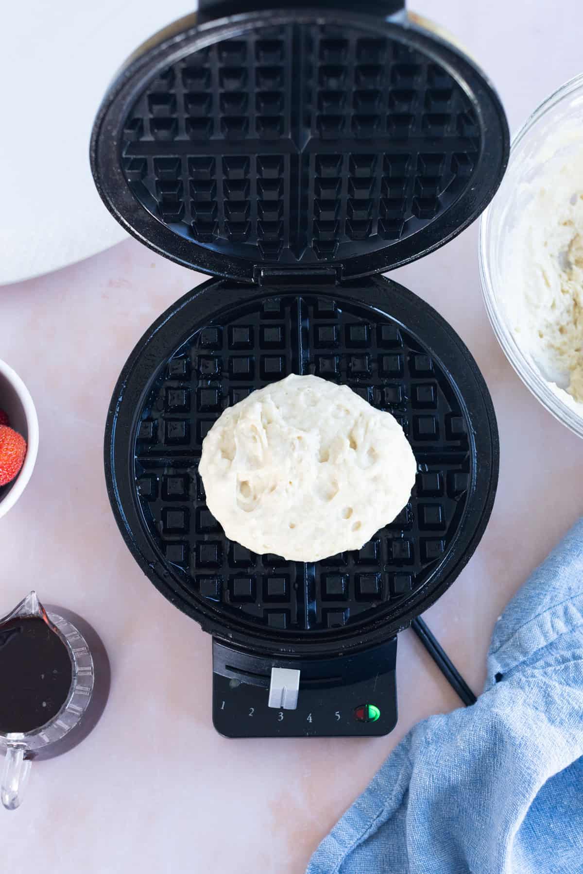 A portion of easy waffle batter sits on an open waffle maker, surrounded by a bowl of batter, strawberries, syrup, and a blue cloth.