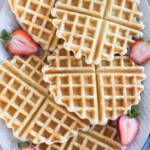 A plate of easy waffles, perfectly round and arranged with sliced strawberries, sits on a white surface.