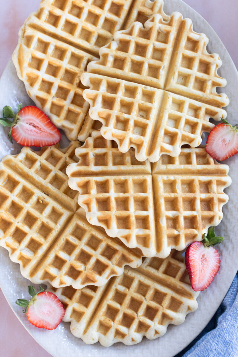 A plate of easy waffles, perfectly round and arranged with sliced strawberries, sits on a white surface.