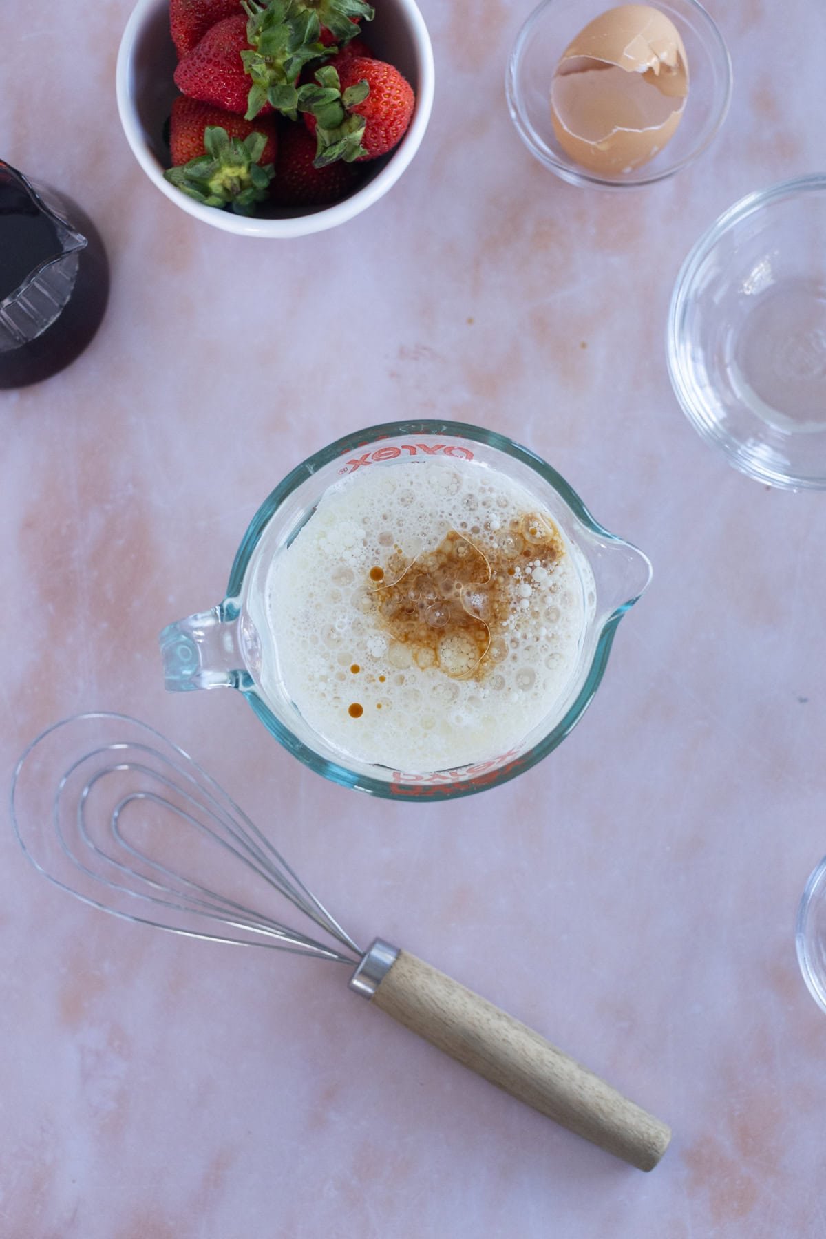 A glass measuring cup with a frothy mixture for easy waffles and brown liquid, surrounded by a whisk, an eggshell, a bowl of strawberries, and an empty glass bowl on a light countertop.