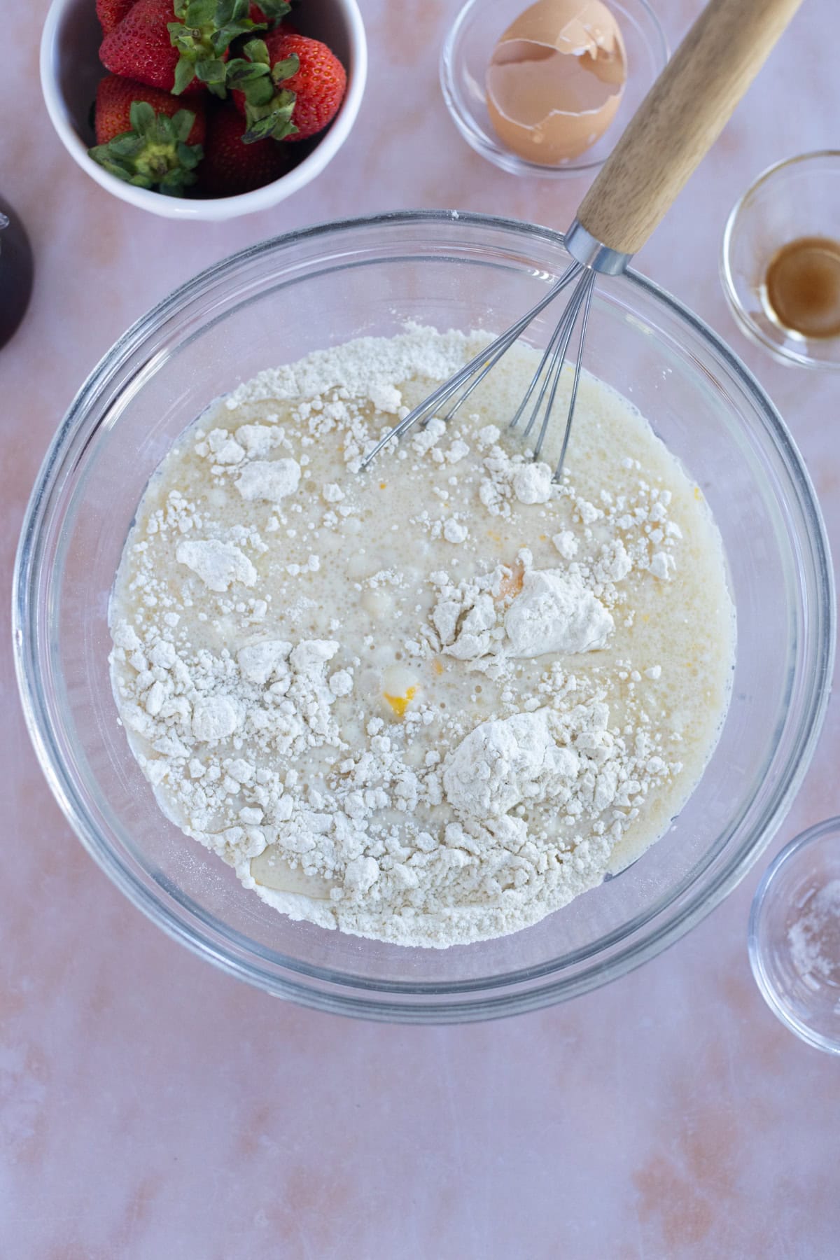 A mixing bowl with flour, milk, and egg being whisked for easy waffles; nearby are a bowl of strawberries, an eggshell, vanilla extract, and an empty glass bowl.
