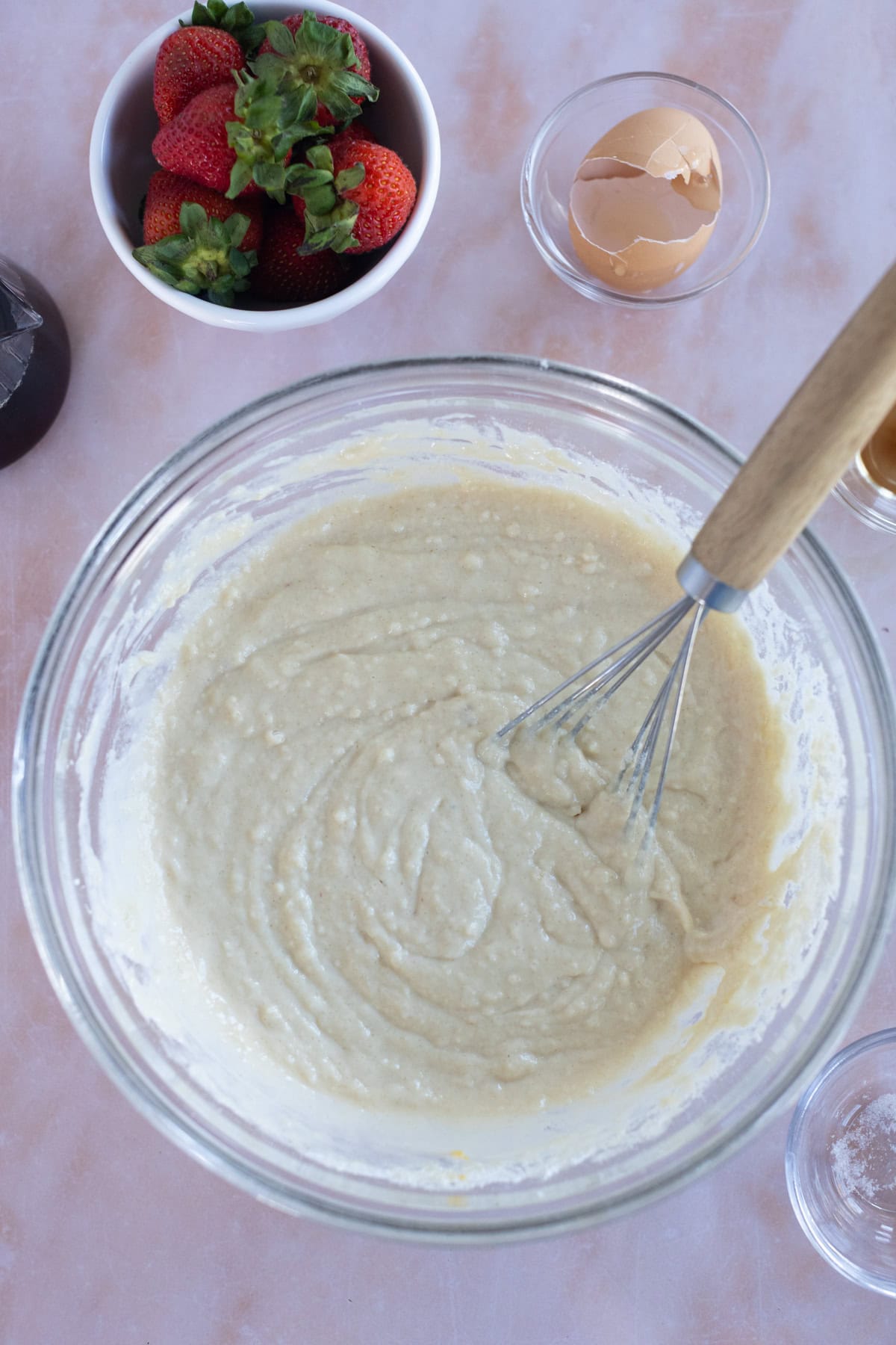 A glass bowl of batter being whisked for easy waffles, with a bowl of strawberries, an eggshell, and small glass bowls on a marble countertop.